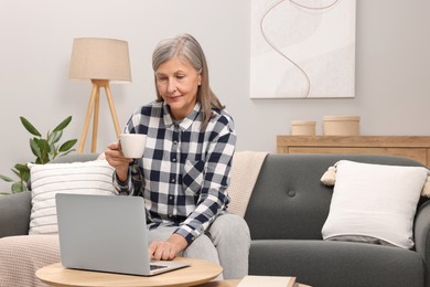 Photo of Beautiful senior woman with cup of drink using laptop at home