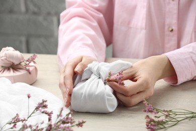 Photo of Furoshiki technique. Woman decorating gift wrapped in white fabric with beautiful pink flower at wooden table, closeup