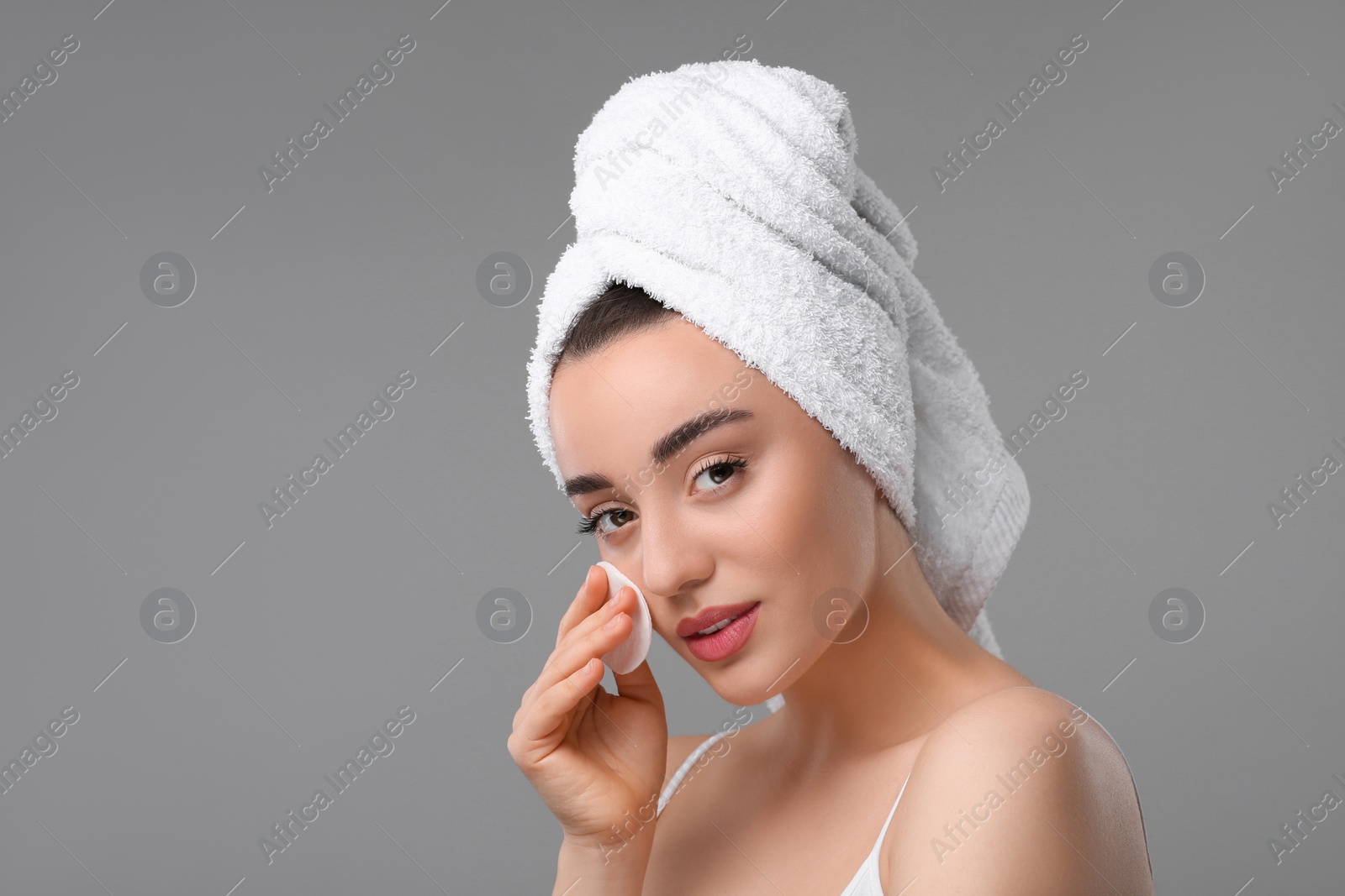 Photo of Beautiful woman in terry towel removing makeup with cotton pad on gray background, space for text