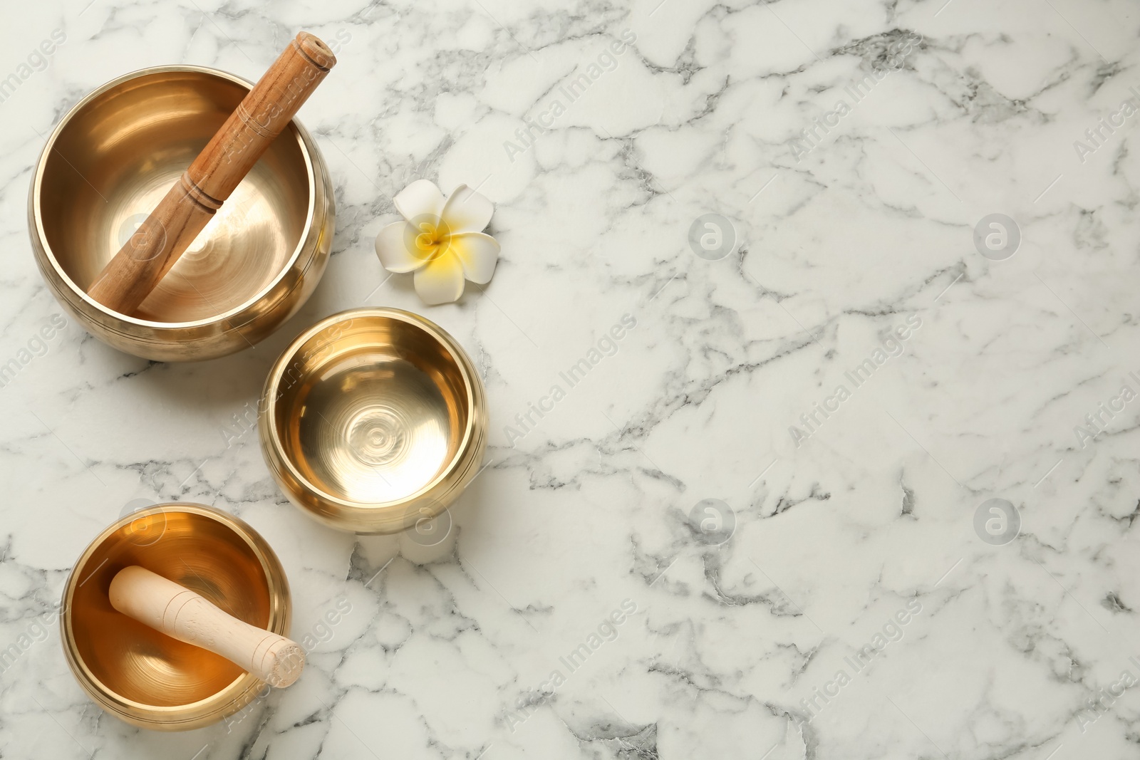 Photo of Golden singing bowls, mallets and flower on white marble table, flat lay. Space for text