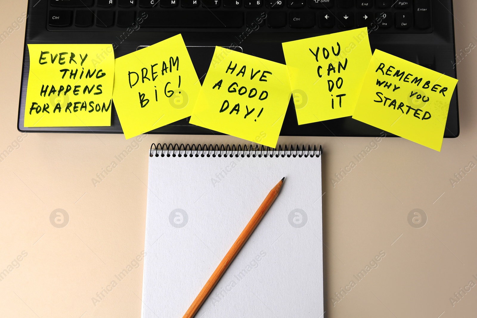 Photo of Laptop with paper notes of life-affirming phrases and notebook on beige desk, flat lay