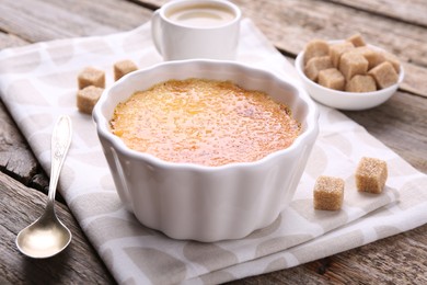 Photo of Delicious creme brulee in bowl, sugar cubes and spoon on wooden table, closeup