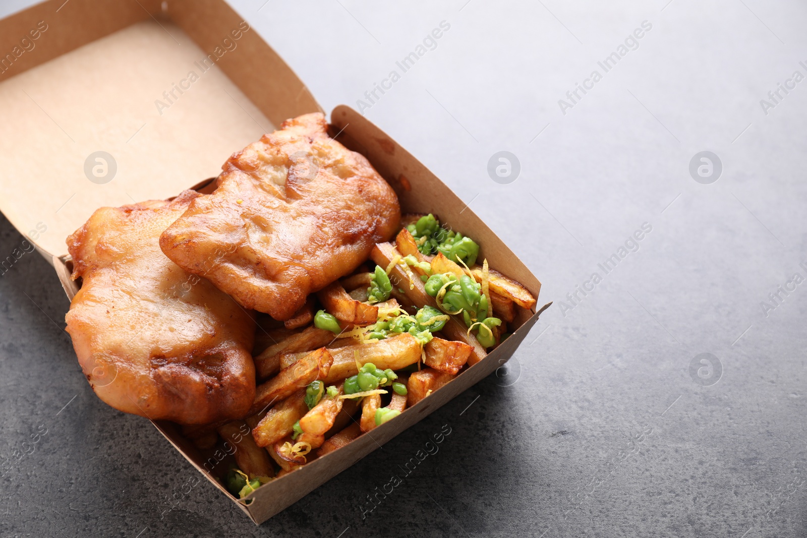 Photo of Tasty fish, chips and peas in paper box on grey table, closeup. Space for text