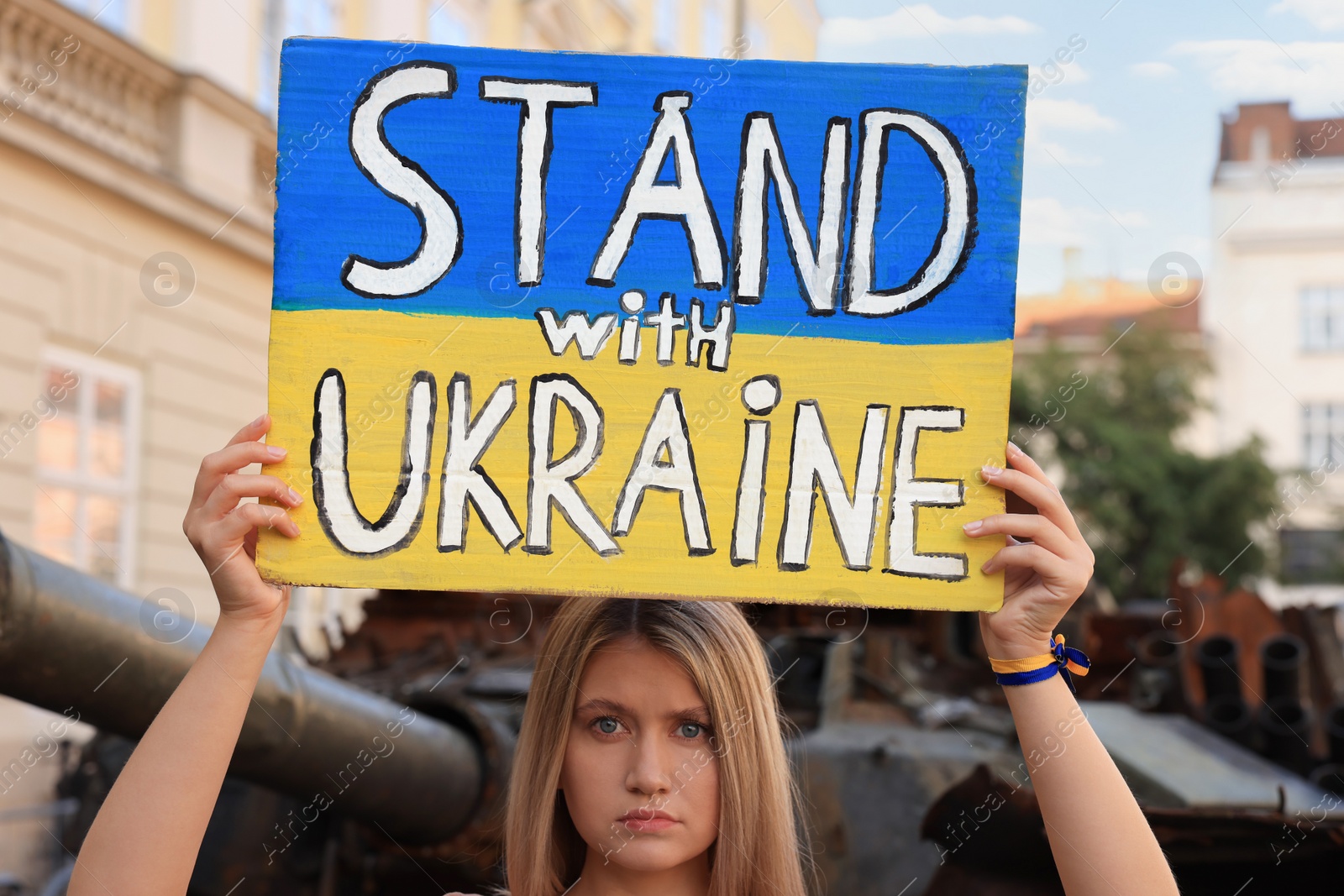 Photo of Sad woman holding poster in colors of national flag with words Stand with Ukraine near broken tank on city street