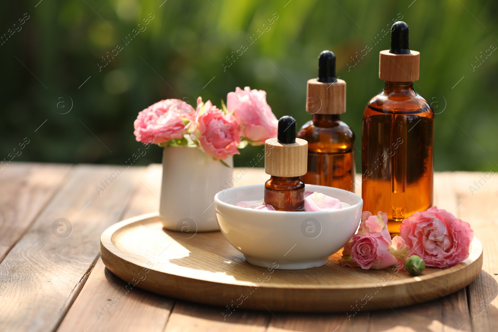 Photo of Bottles of rose essential oil and flowers on wooden table outdoors