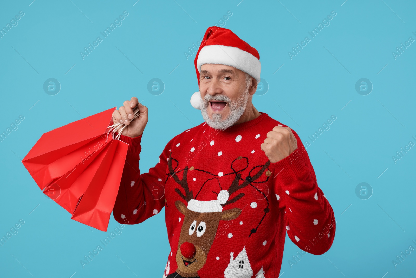 Photo of Happy senior man in Christmas sweater and Santa hat with shopping bags on light blue background