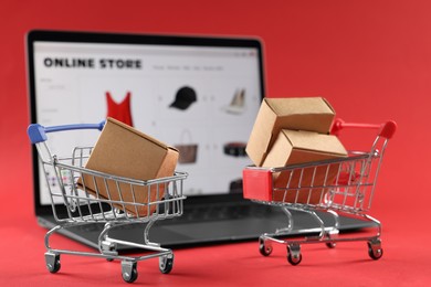 Photo of Online store. Mini shopping carts, parcels and laptop on red background, selective focus