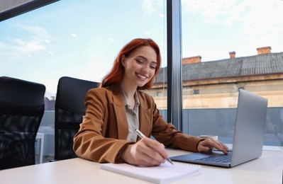 Happy woman taking notes while working with laptop at white desk in office