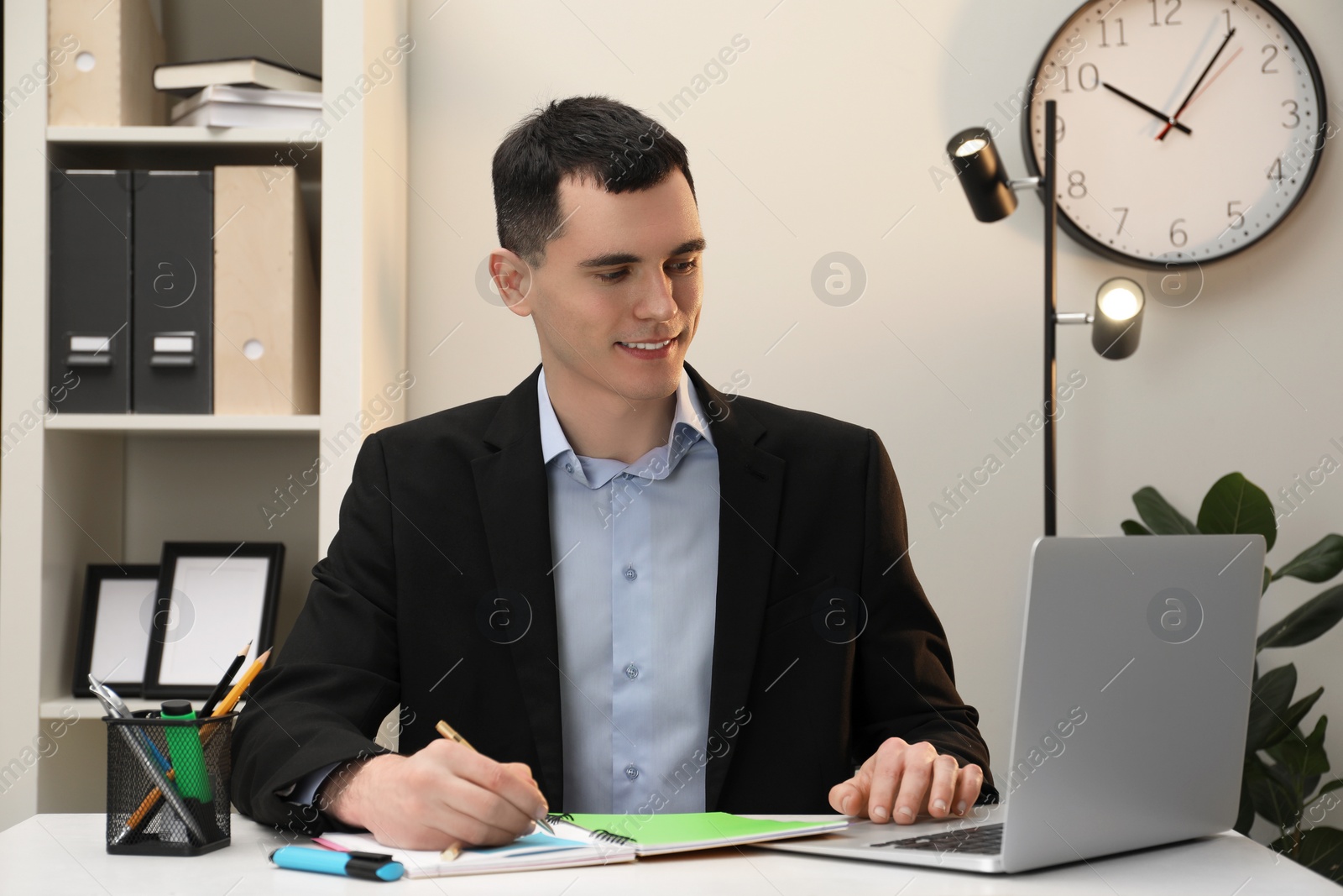 Photo of Man taking notes while using laptop at table in office