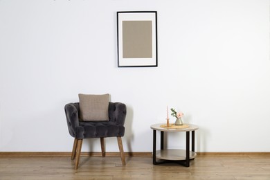 Photo of Comfortable armchair, cushion, table and decor elements in room with white wall. Interior design