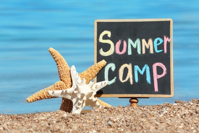 Photo of Little blackboard with text SUMMER CAMP and sea stars on sand near sea