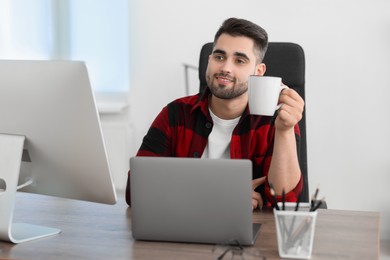 Photo of Happy young programmer drinking tea while working with laptop in office