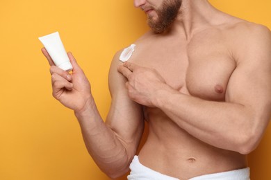 Man applying moisturizing cream onto his shoulder on orange background, closeup