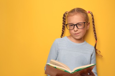 Photo of Cute little girl with glasses and textbook on yellow background. Space for text