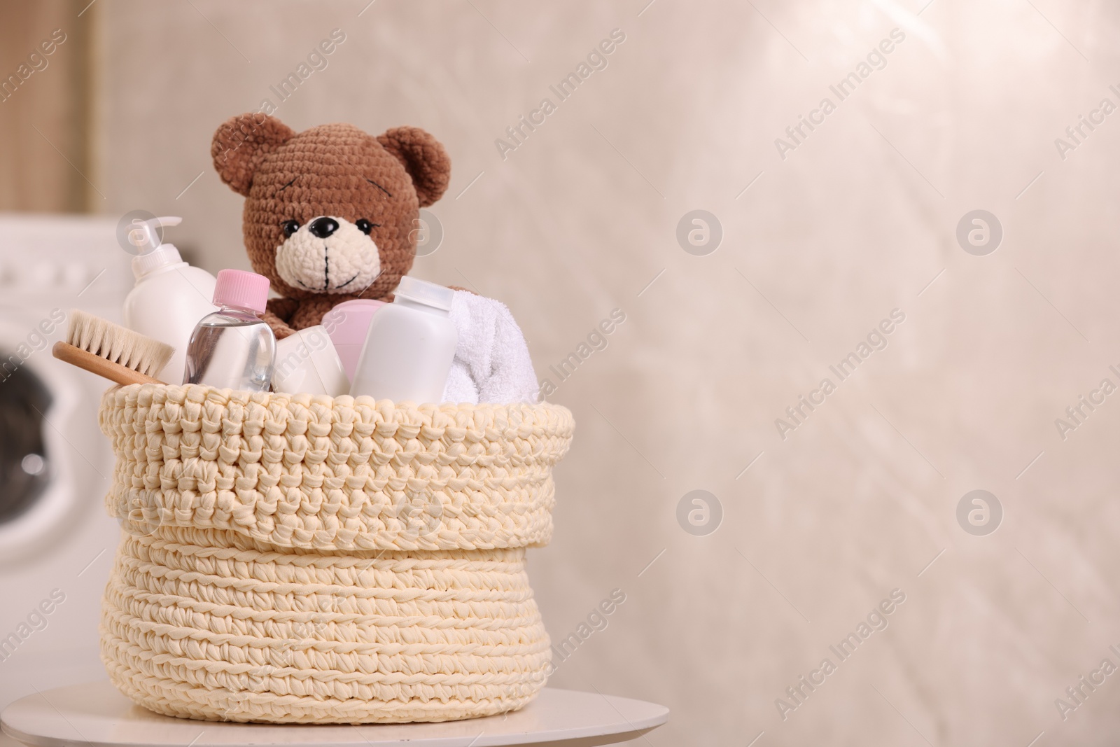 Photo of Knitted basket with baby cosmetic products, bath accessories and toy bear on white table indoors. Space for text