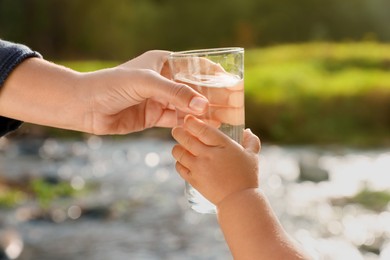 Mother giving her daughter glass of fresh water near stream on sunny day, closeup