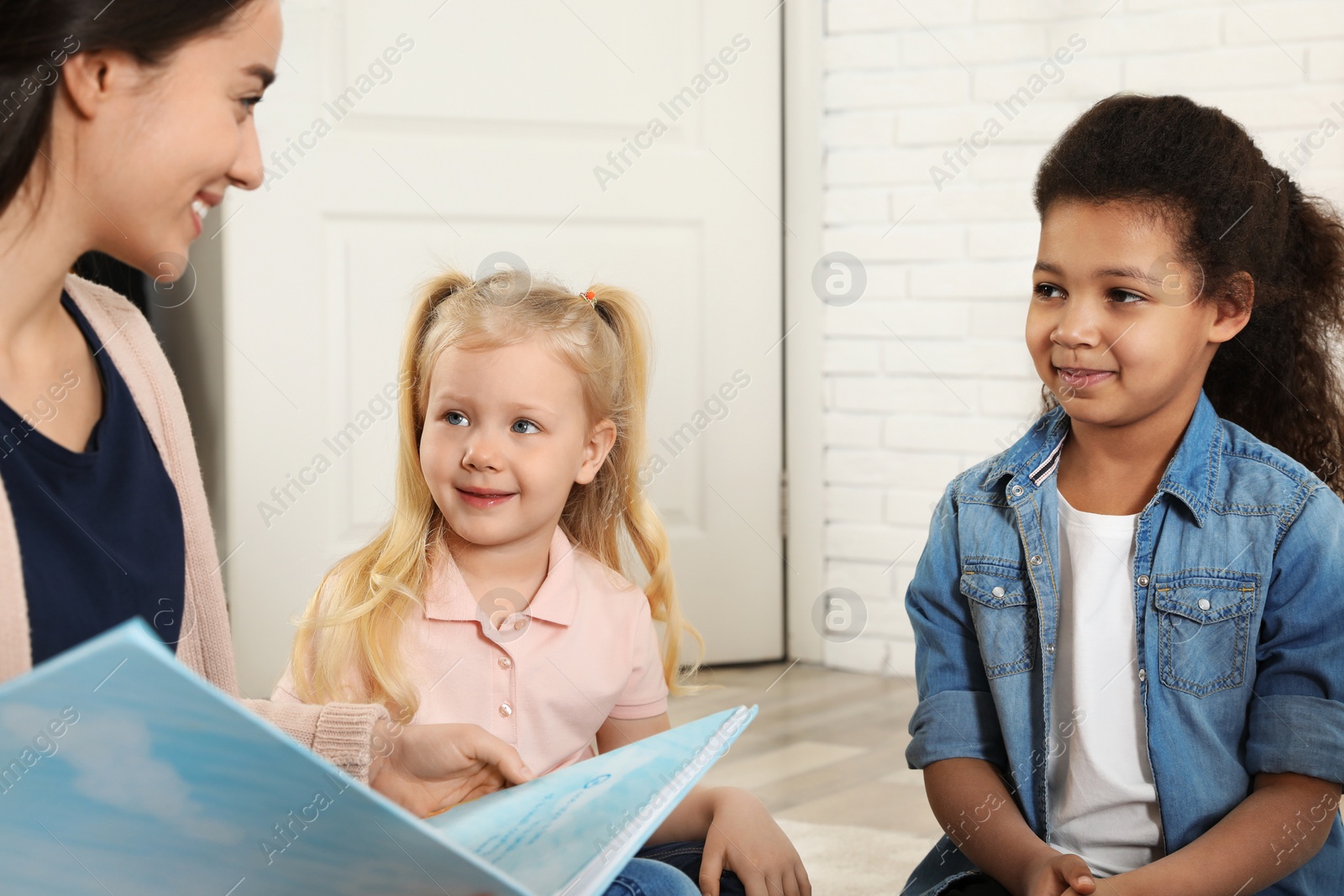 Photo of Kindergarten teacher reading book to cute little children indoors