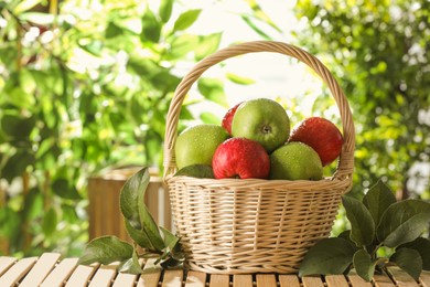 Photo of Different wet apples in wicker basket and green leaves on wooden table outdoors