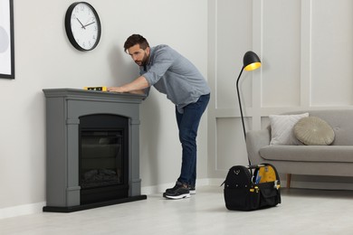 Photo of Man using construction level for installing electric fireplace near wall in room