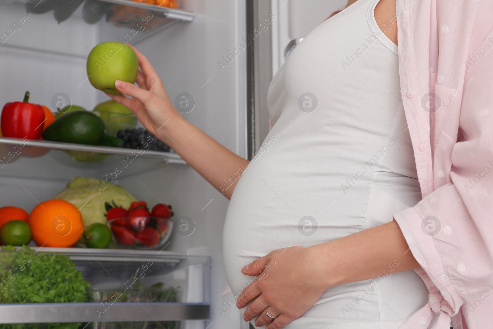 Photo of Young pregnant woman with apple near fridge at home, closeup. Healthy eating