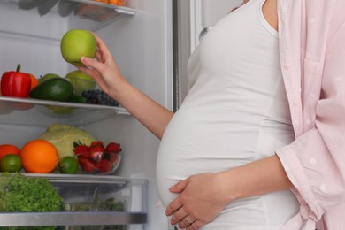 Photo of Young pregnant woman with apple near fridge at home, closeup. Healthy eating