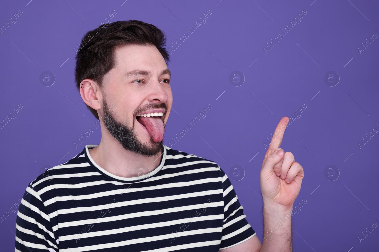 Photo of Happy man showing his tongue and pointing on purple background. Space for text