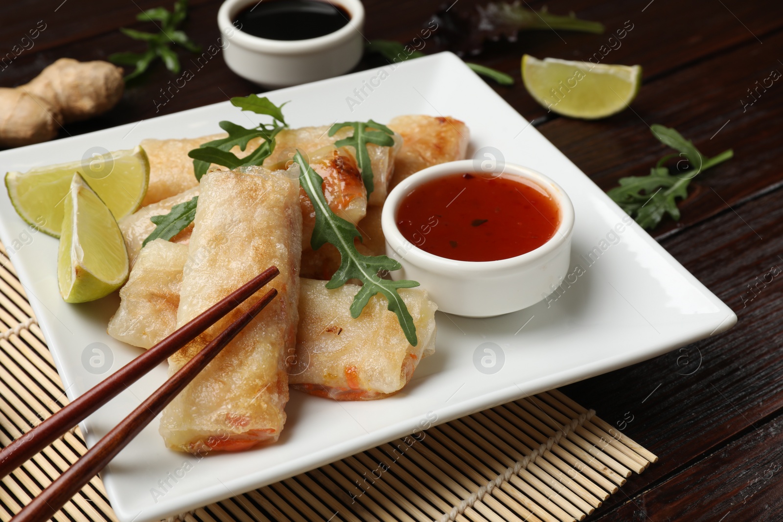 Photo of Tasty fried spring rolls, arugula, lime and sauce served on wooden table, closeup