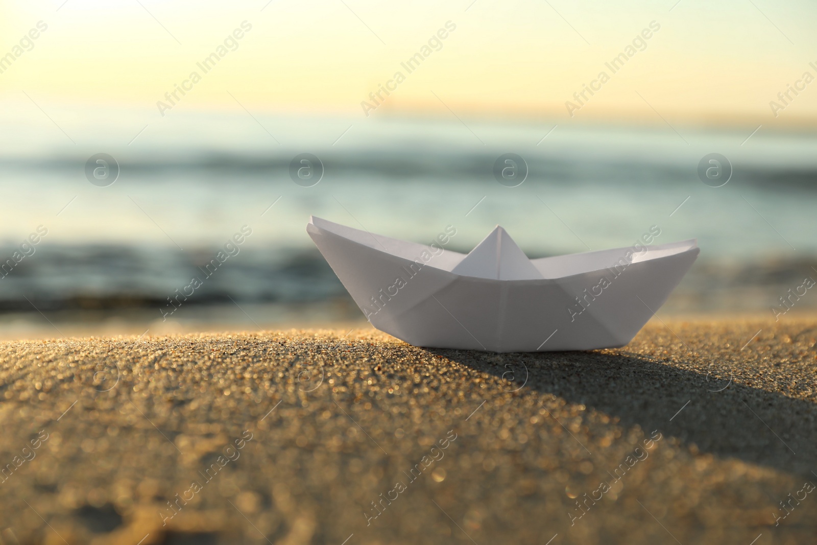 Photo of White paper boat on sand near sea at sunset, closeup. Space for text