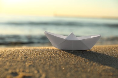 Photo of White paper boat on sand near sea at sunset, closeup. Space for text