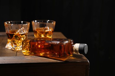 Whiskey with ice cubes in glasses and bottle on wooden crate against black background, closeup