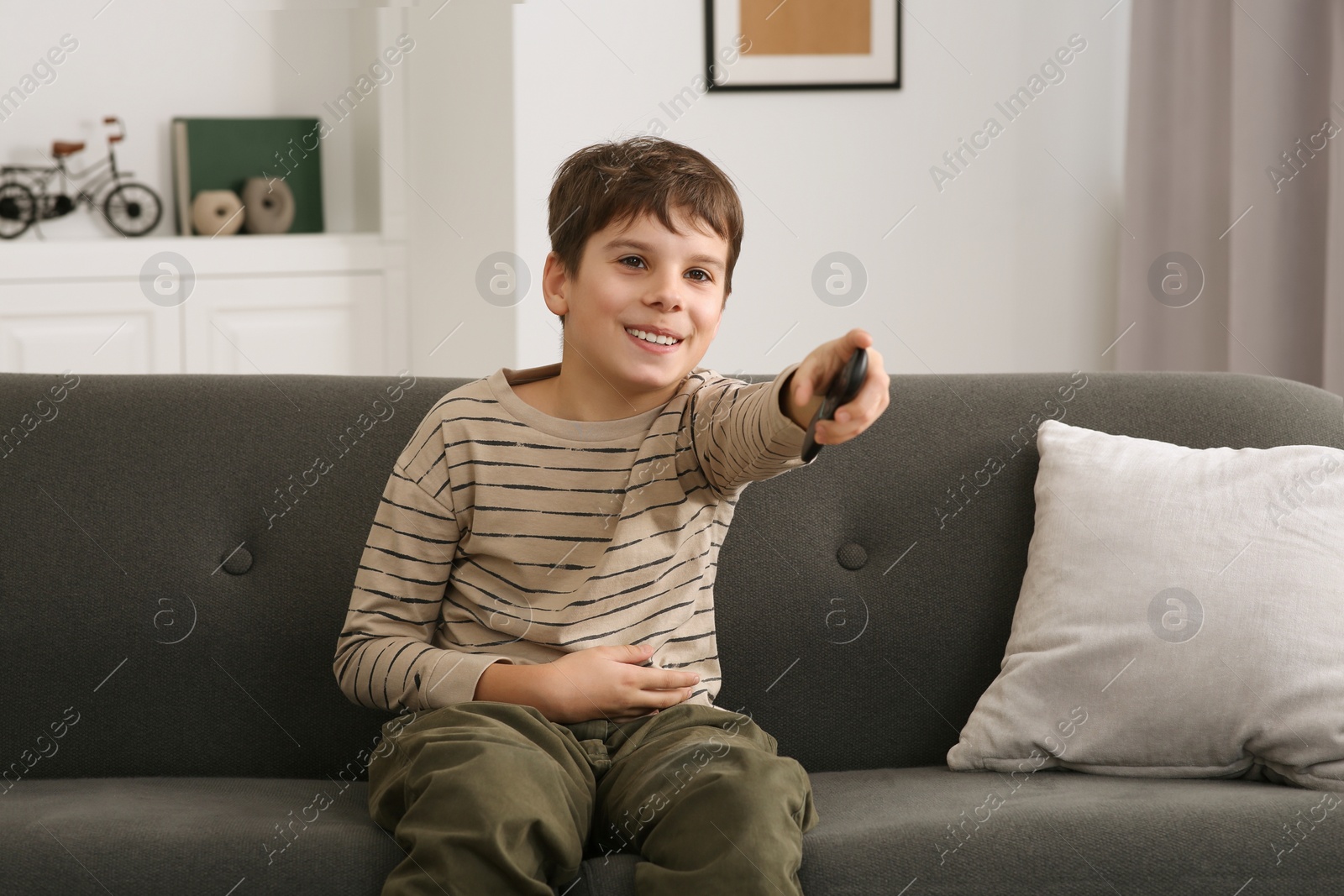 Photo of Happy boy changing TV channels with remote control on sofa at home