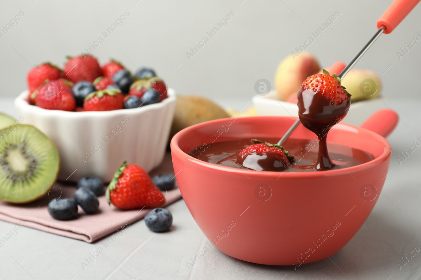 Photo of Dipping strawberry into fondue pot with chocolate on grey table, closeup