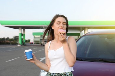 Beautiful young woman with coffee eating doughnut near car at gas station