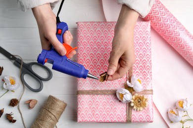 Photo of Woman using hot glue gun to decorate gift at white wooden table, top view