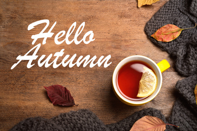 Text Hello Autumn, cup of tea, scarf and dry leaves on wooden background, top view