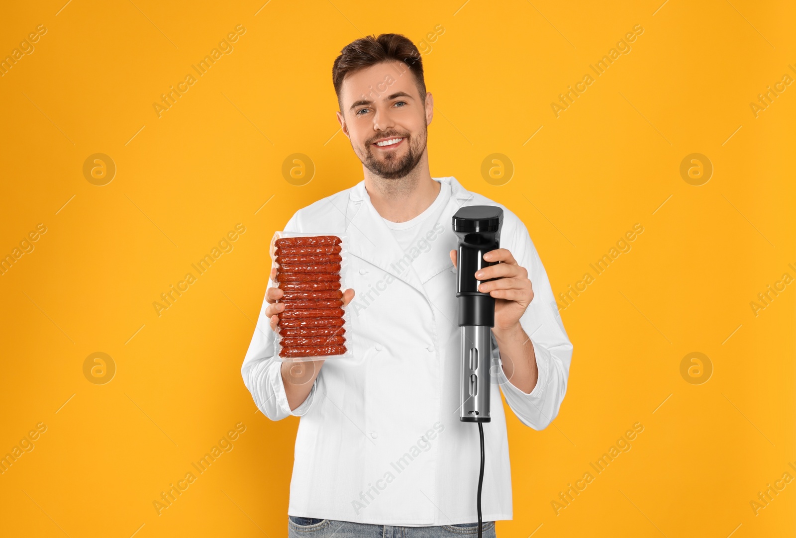 Photo of Smiling chef holding sous vide cooker and sausages in vacuum pack on orange background