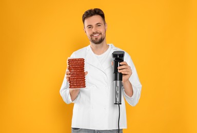 Photo of Smiling chef holding sous vide cooker and sausages in vacuum pack on orange background