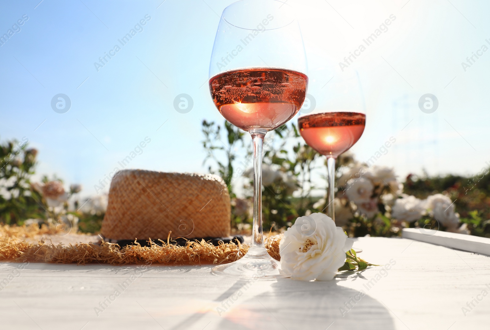 Photo of Glasses of rose wine, straw hat and beautiful flower on white wooden table outdoors. Space for text