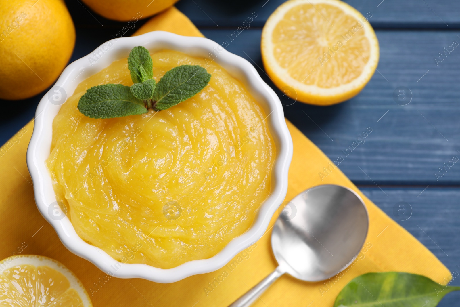 Photo of Delicious lemon curd in bowl, fresh citrus fruits and spoon on blue wooden table, flat lay