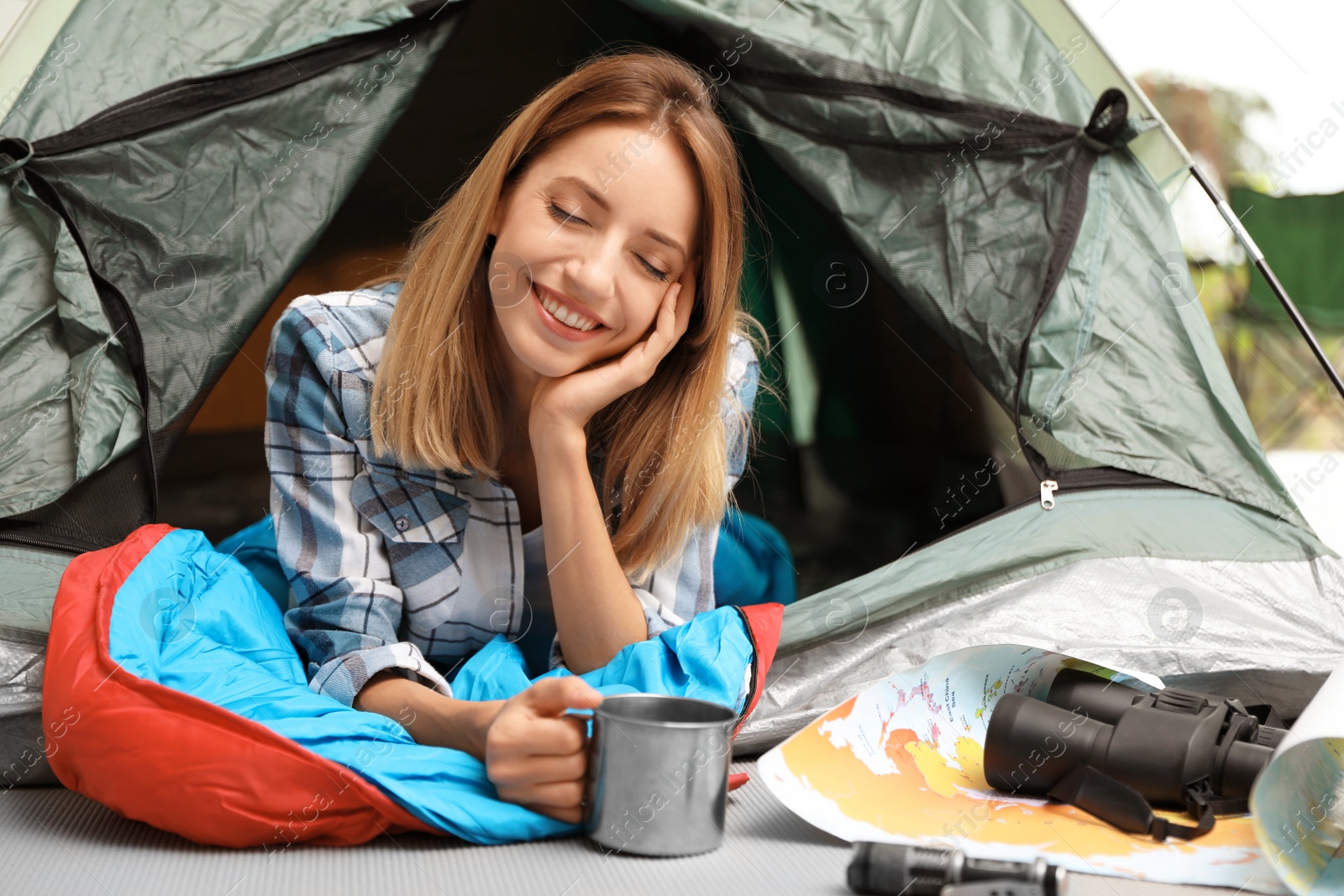 Photo of Young woman in sleeping bag with mug looking outside of tent