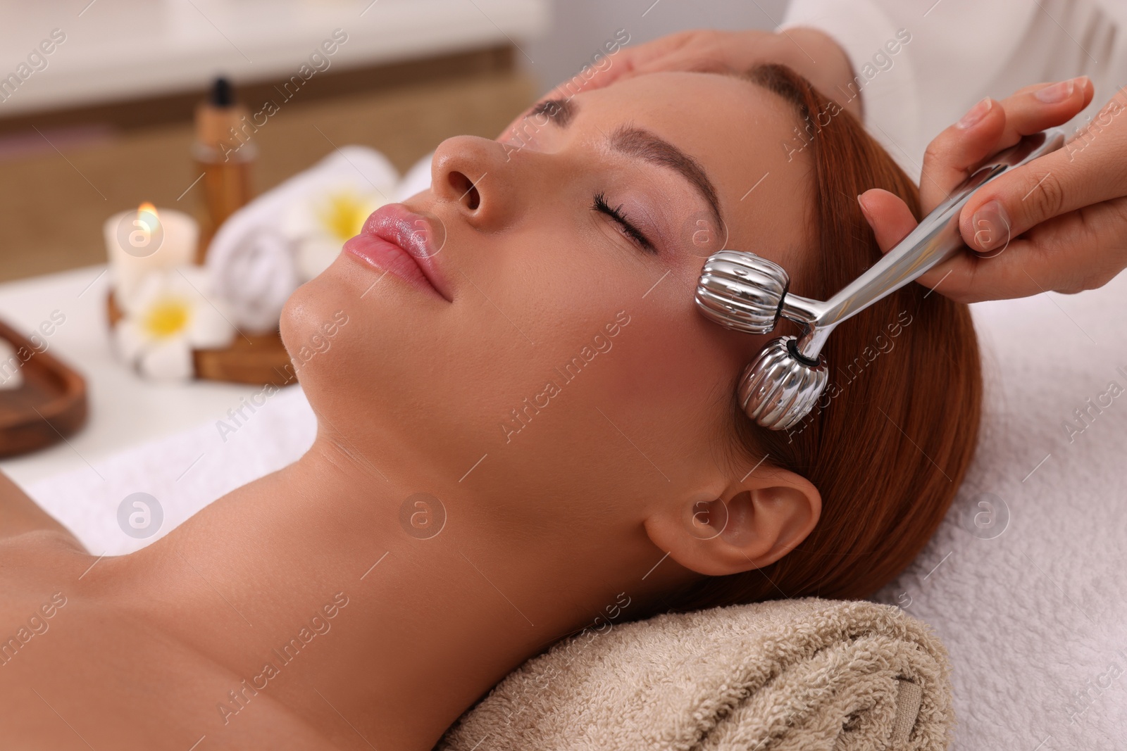 Photo of Young woman receiving facial massage with metal roller in beauty salon, closeup