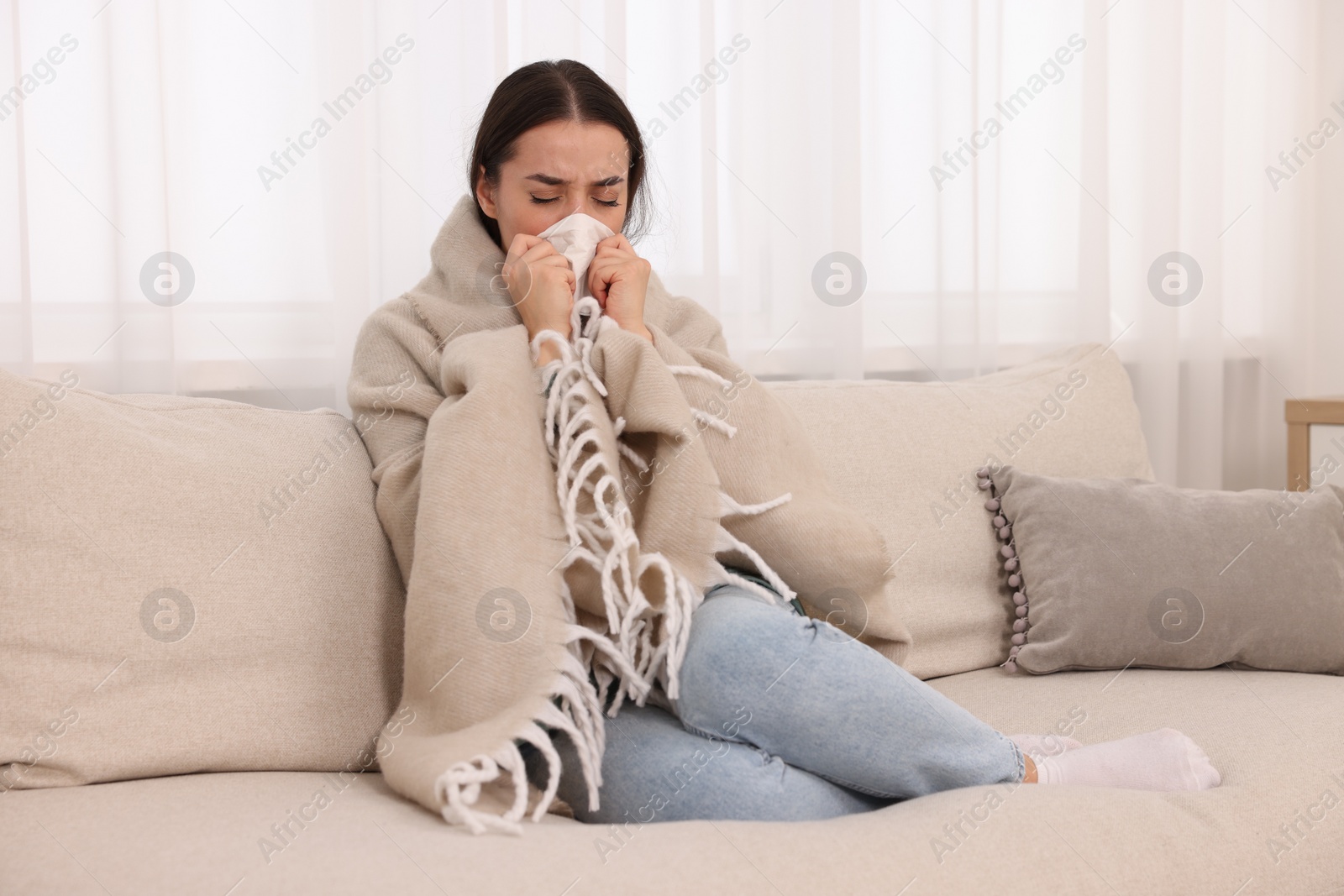 Photo of Sick woman wrapped in blanket with tissue blowing nose on sofa at home. Cold symptoms