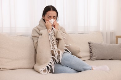 Sick woman wrapped in blanket with tissue blowing nose on sofa at home. Cold symptoms