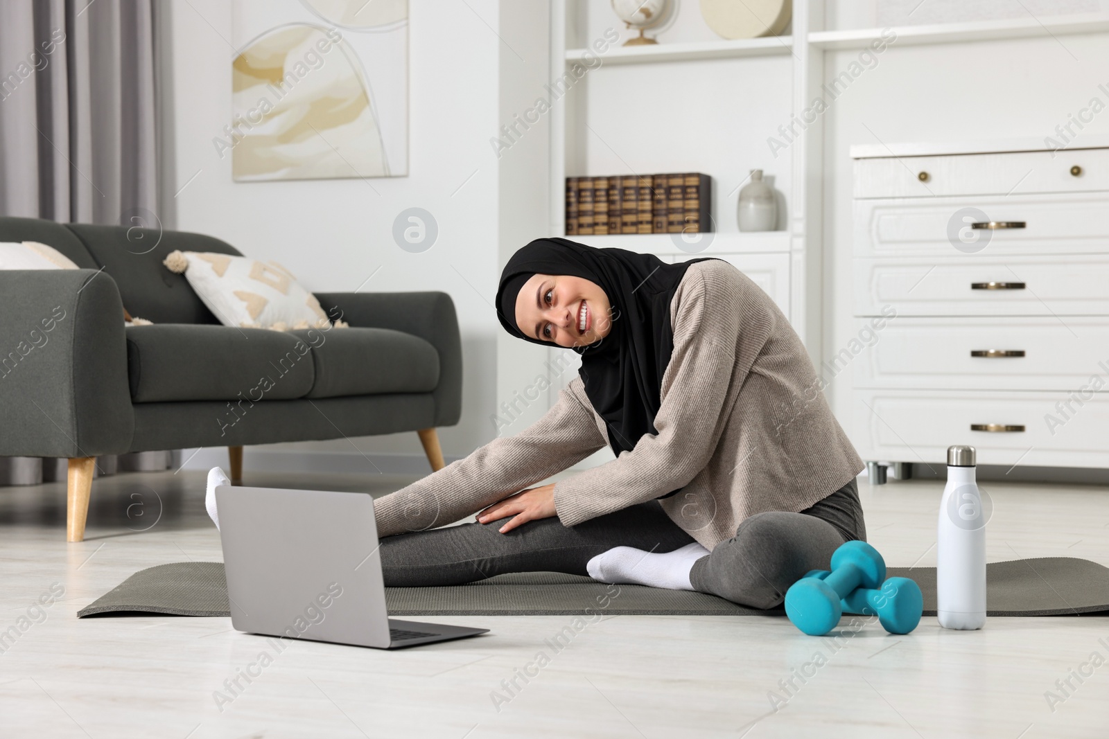 Photo of Muslim woman in hijab stretching near laptop on fitness mat at home