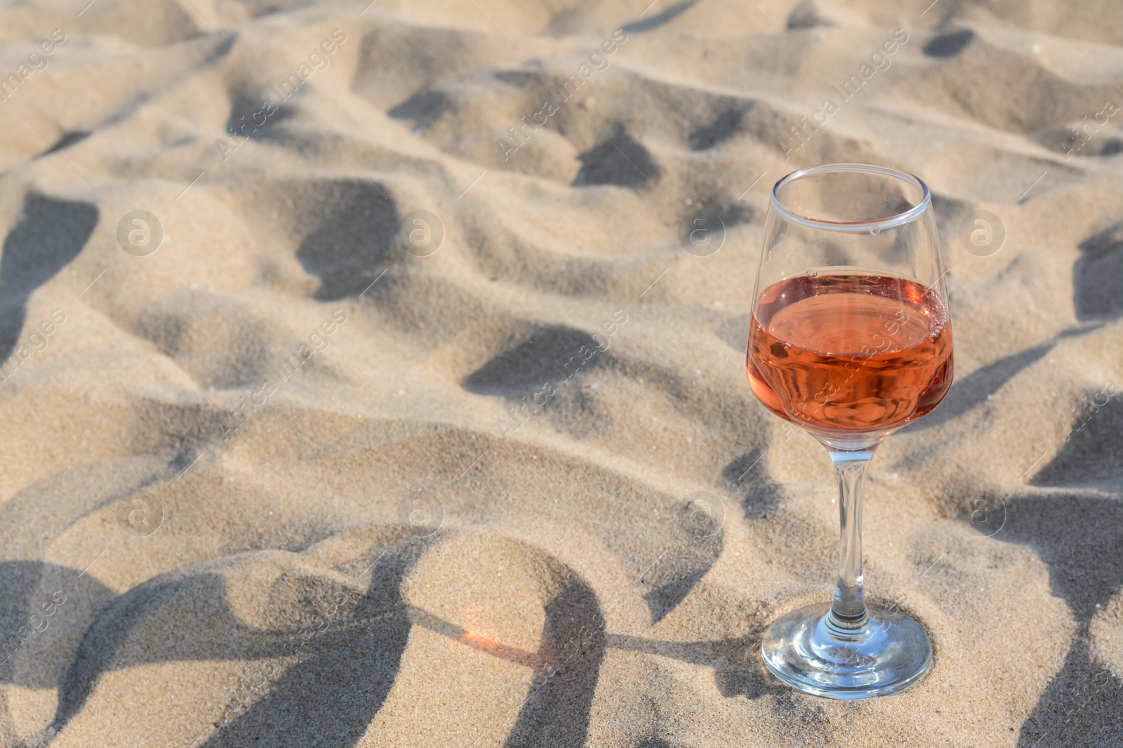 Photo of Glass of tasty rose wine on sand, space for text