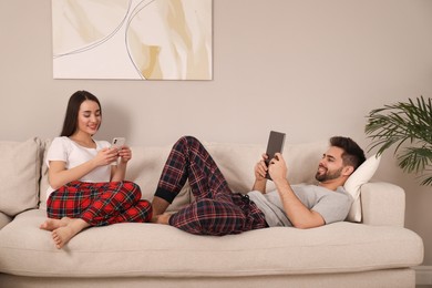 Happy couple in pajamas with gadgets on sofa at home