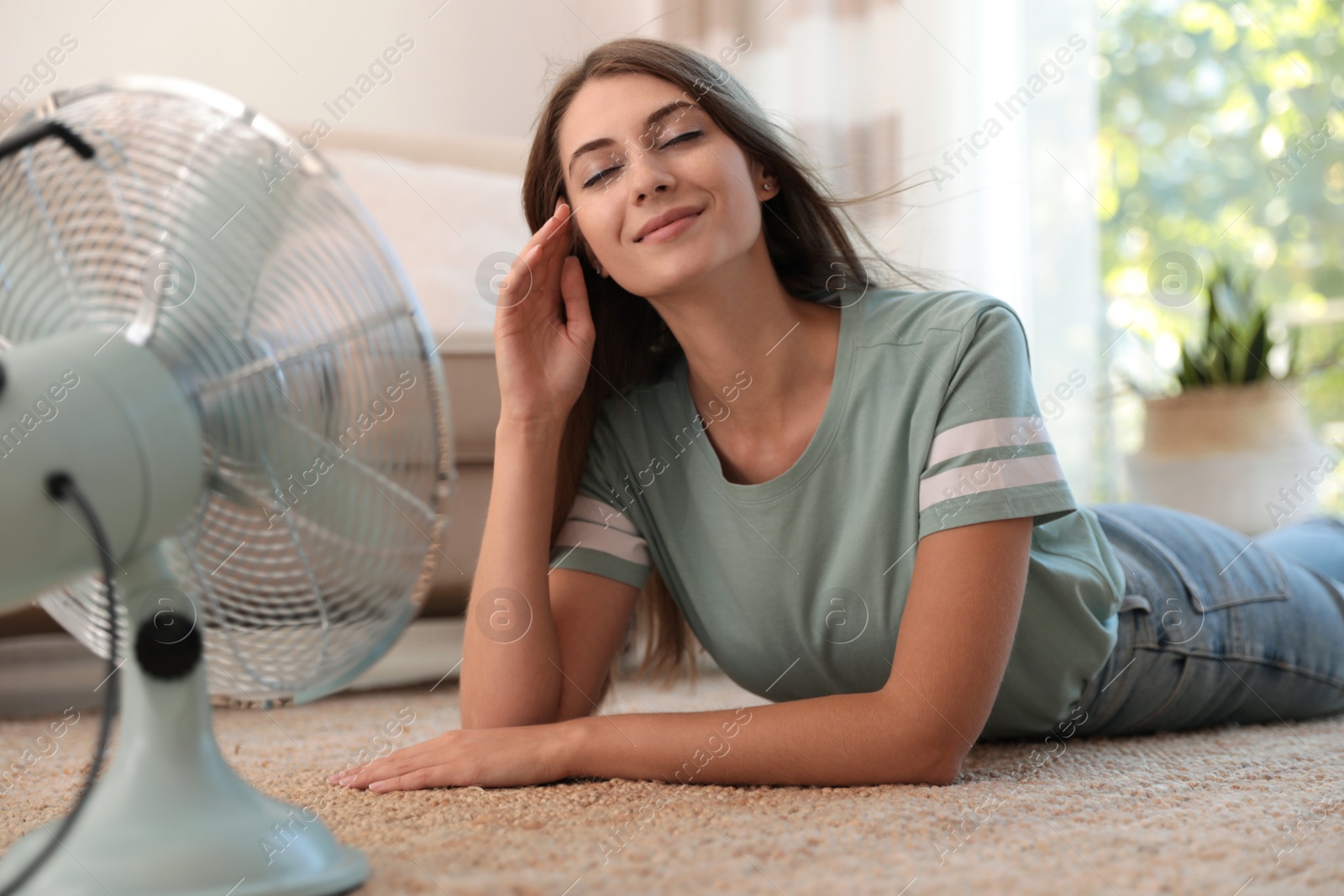 Photo of Woman enjoying air flow from fan on floor in living room. Summer heat