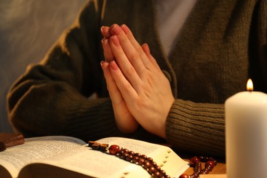 Woman praying at table with burning candle and Bible, closeup