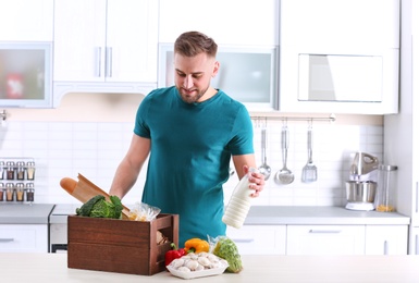 Photo of Young man with wooden crate full of products at table in kitchen. Food delivery service