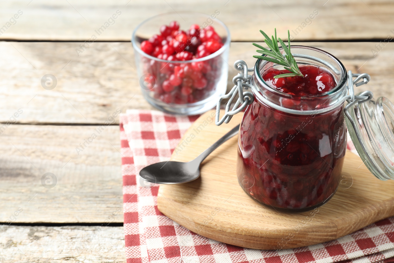 Photo of Fresh cranberry sauce in glass jar, rosemary and spoon on light wooden table, space for text